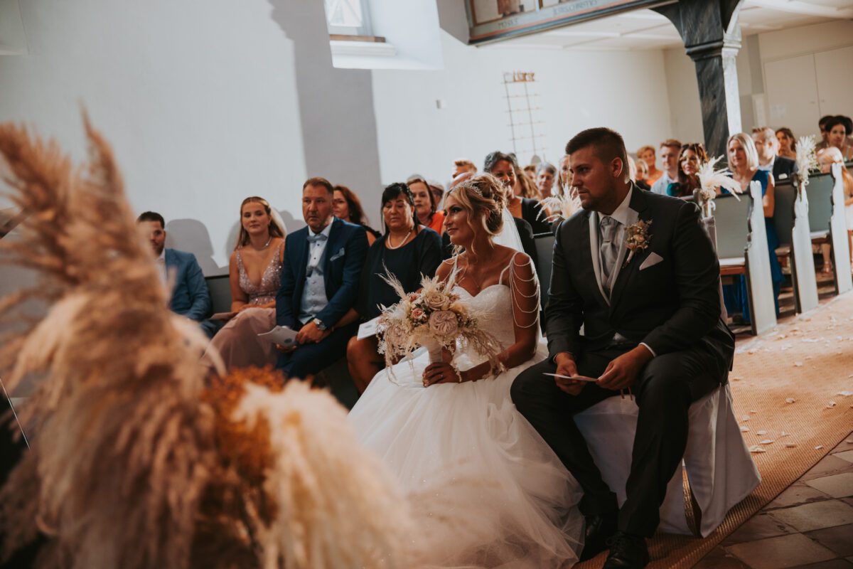 Braut und Bräutigam sitzen während ihrer Hochzeitszeremonie in einer mit Gästen gefüllten Kirche in Frankfurt nebeneinander. Die Braut hält einen Blumenstrauß in der Hand und trägt ein weißes Kleid, während der Bräutigam einen dunklen Anzug trägt. Sonnenlicht fällt durch die Fenster und wirft einen sanften Schein auf das Paar und seine Gäste – perfekt für ein Fotoshooting. - Elis Wedding - Hochzeitsfotograf Frankfurt