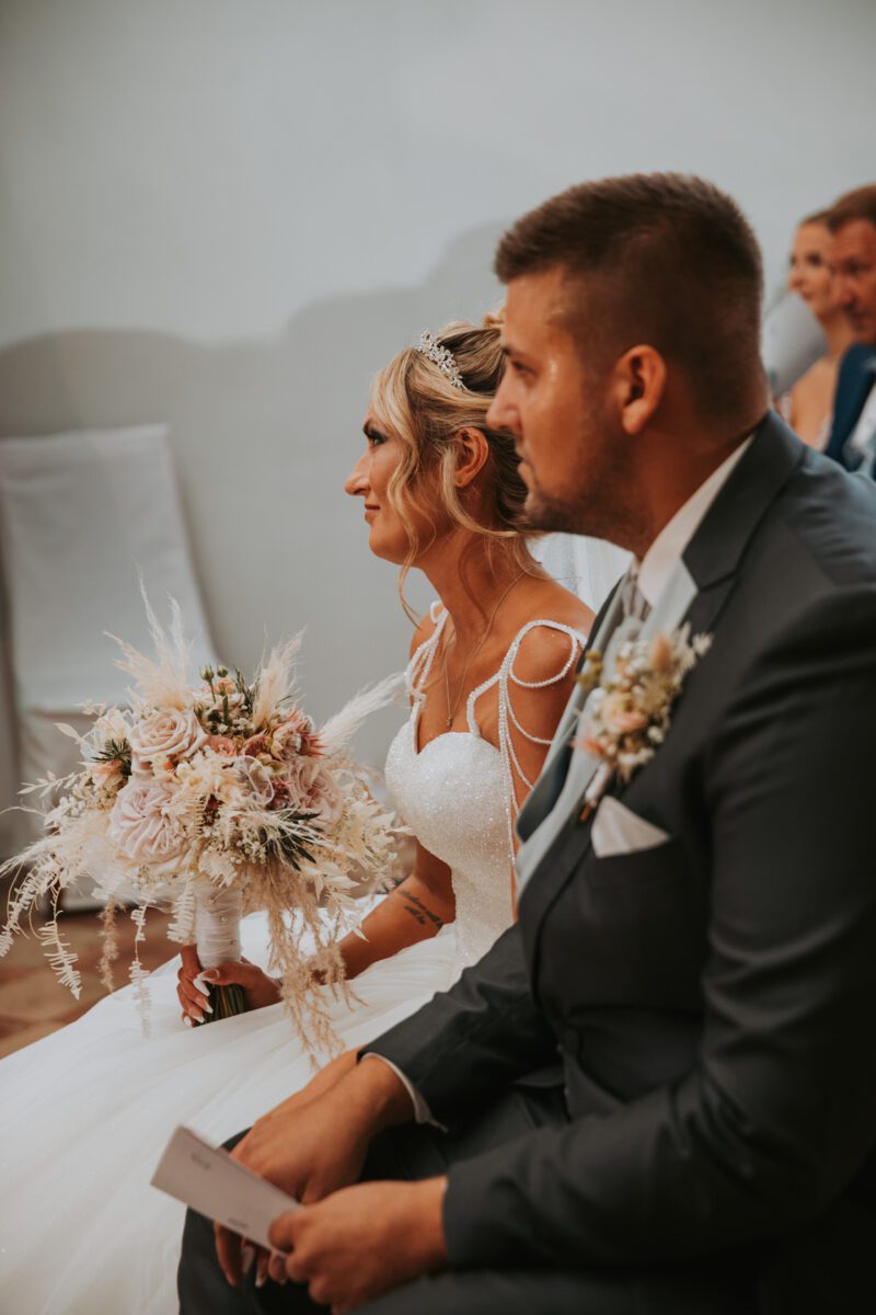 Braut und Bräutigam sitzen an ihrem Hochzeitstag drinnen. Die Braut, gekleidet in ein weißes Kleid mit einem leichten, aufwendigen Blumenstrauß, blickt nach vorne. Der Bräutigam, der einen dunklen Anzug mit einer Ansteckblume trägt, blickt während ihrer standesamtlichen Trauung ebenfalls nach vorne. Beide wirken aufmerksam, während die Gäste hinter ihnen sitzen. - Elis Wedding - Hochzeitsfotograf Frankfurt