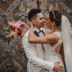 Ein Paar in Hochzeitskleidung teilt einen intimen Moment nach seiner standesamtlichen Trauung. Der Bräutigam, der einen weißen Anzug und eine schwarze Fliege trägt, berührt sanft das Gesicht der Braut. Die Braut, in einem Spitzenhochzeitskleid mit offenem Rücken und langem Schleier, hält einen Strauß rosa Blumen. Sie stehen vor einer großen, kunstvoll gestalteten Holztür in Frankfurt. - Elis Wedding - Hochzeitsfotograf Frankfurt