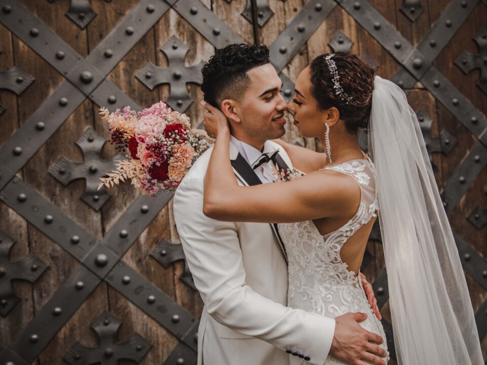 Ein Paar in Hochzeitskleidung teilt einen intimen Moment nach seiner standesamtlichen Trauung. Der Bräutigam, der einen weißen Anzug und eine schwarze Fliege trägt, berührt sanft das Gesicht der Braut. Die Braut, in einem Spitzenhochzeitskleid mit offenem Rücken und langem Schleier, hält einen Strauß rosa Blumen. Sie stehen vor einer großen, kunstvoll gestalteten Holztür in Frankfurt. - Elis Wedding - Hochzeitsfotograf Frankfurt
