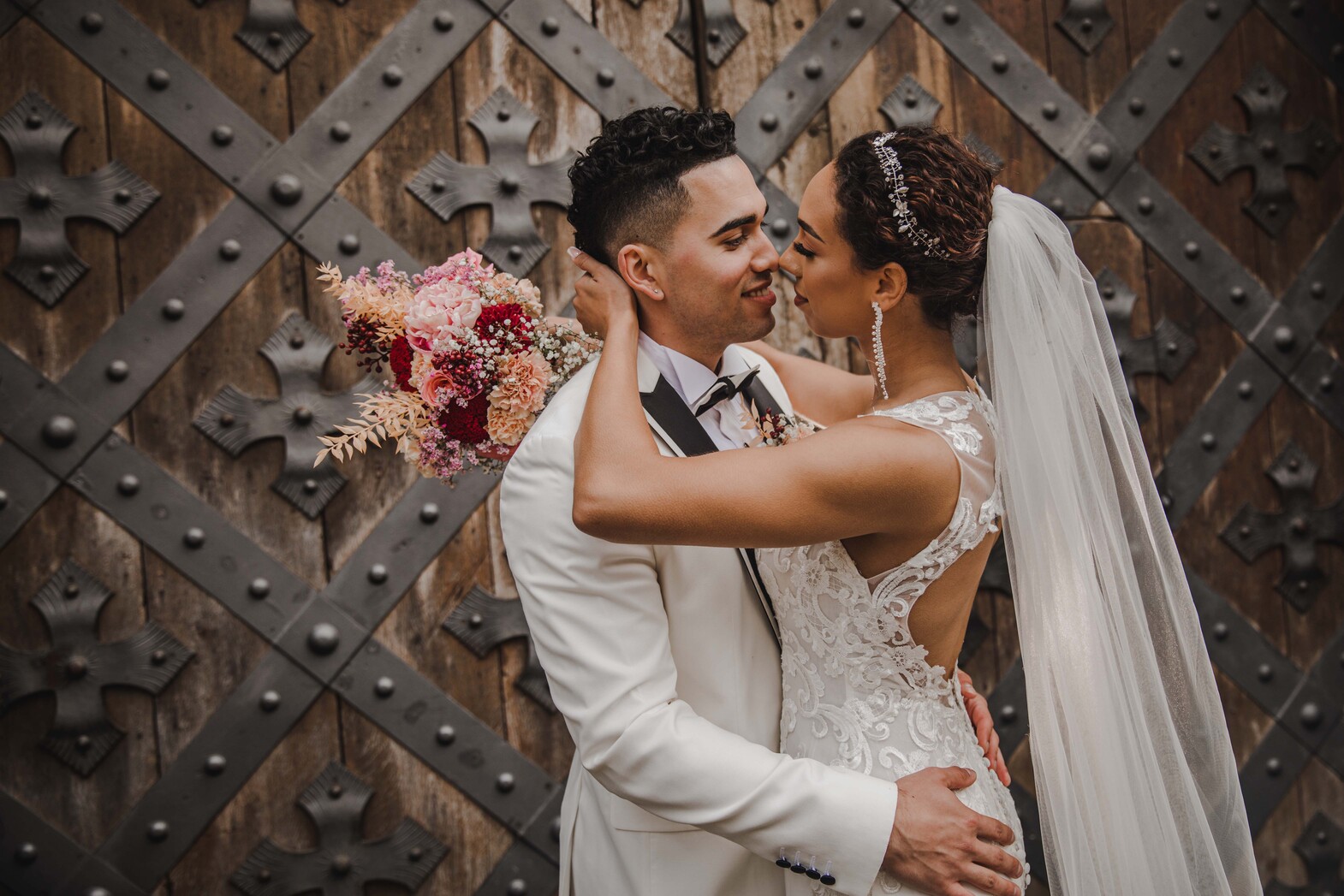 Ein Paar in Hochzeitskleidung teilt einen intimen Moment nach seiner standesamtlichen Trauung. Der Bräutigam, der einen weißen Anzug und eine schwarze Fliege trägt, berührt sanft das Gesicht der Braut. Die Braut, in einem Spitzenhochzeitskleid mit offenem Rücken und langem Schleier, hält einen Strauß rosa Blumen. Sie stehen vor einer großen, kunstvoll gestalteten Holztür in Frankfurt. - Elis Wedding - Hochzeitsfotograf Frankfurt