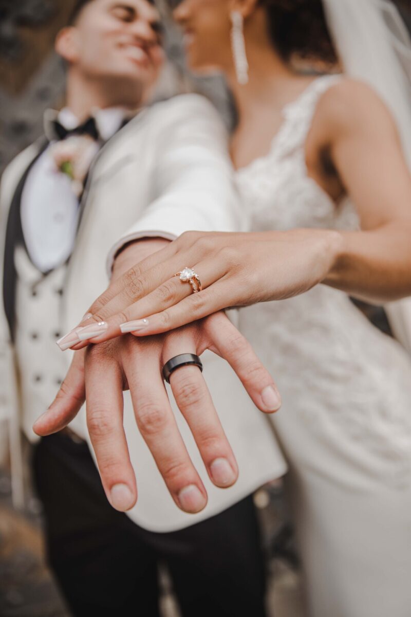 Ein Paar in eleganter Hochzeitskleidung zeigt stolz seine Eheringe während eines Fotoshootings. Der Bräutigam in einem weißen Smoking streckt seine Hand aus, während die Braut in einem Spitzenhochzeitskleid ihre Hand darüberlegt und ihren Solitär-Verlobungsring und ihren Ehering präsentiert. Beide lächeln und schauen sich an. - Elis Wedding - Hochzeitsfotograf Frankfurt