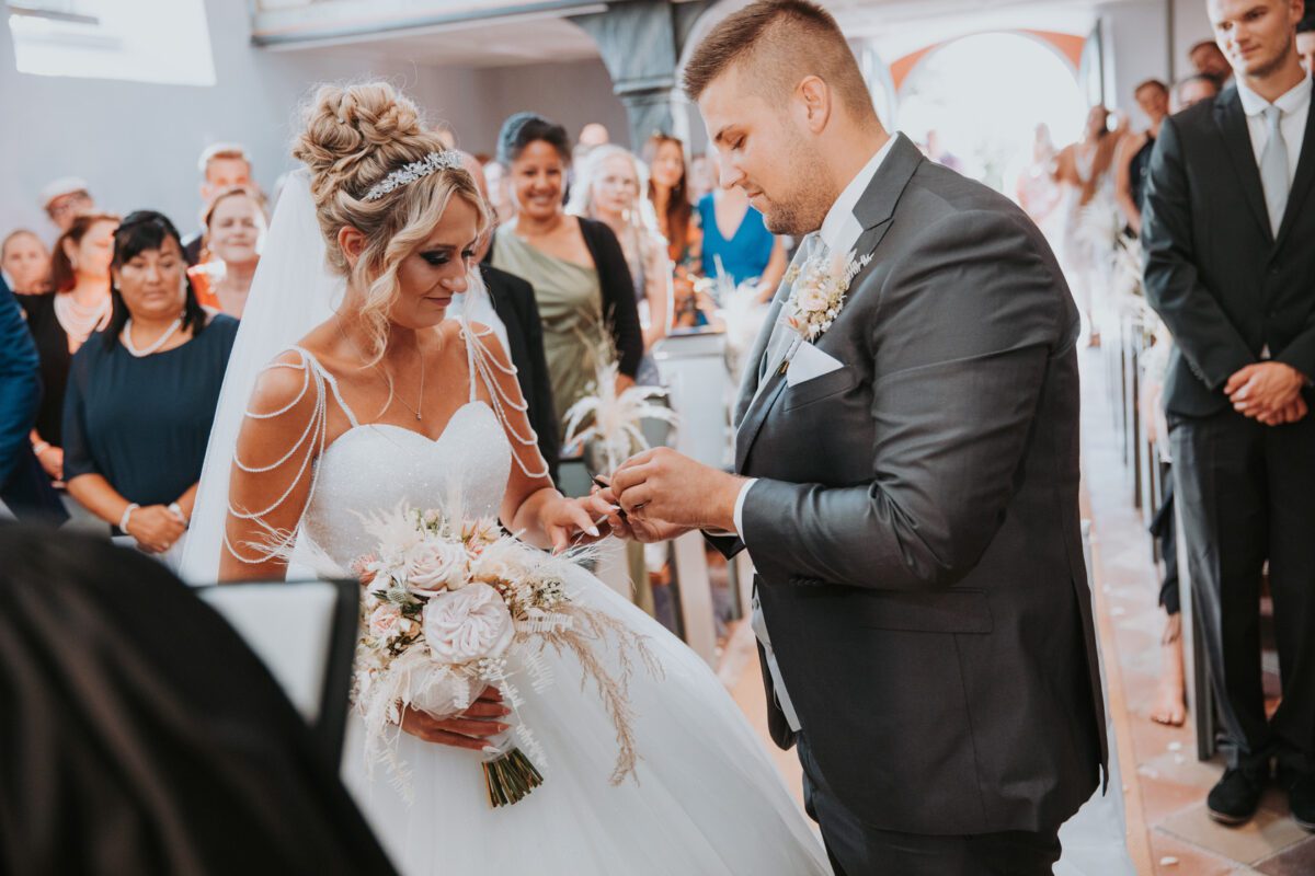 Braut und Bräutigam tauschen während ihrer Hochzeitszeremonie in einer Kirche die Ringe aus. Die Braut, in einem weißen Kleid und mit einem Blumenstrauß in der Hand, sieht zu, wie der Bräutigam, in einem grauen Anzug, ihr einen Ring an den Finger steckt. Ein Hochzeitsfotograf aus Frankfurt fängt den Moment ein, während lächelnde Gäste in den Kirchenbänken dem Paar zuschauen. - Elis Wedding - Hochzeitsfotograf Frankfurt