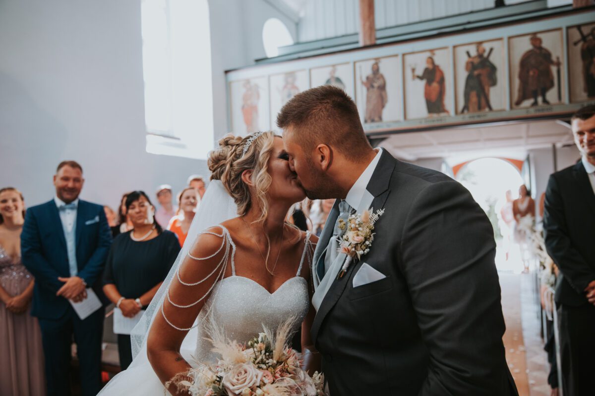 Braut und Bräutigam küssen sich während ihrer standesamtlichen Trauung in einer Kirche. Die Braut trägt ein weißes Kleid mit zarten Trägern und hält einen Blumenstrauß in der Hand, und der Bräutigam trägt einen grauen Anzug mit einer Ansteckblume. Die Gäste stehen im Hintergrund, lächeln und beobachten den Moment, der von ihrem Hochzeitsfotografen festgehalten wurde. - Elis Wedding - Hochzeitsfotograf Frankfurt