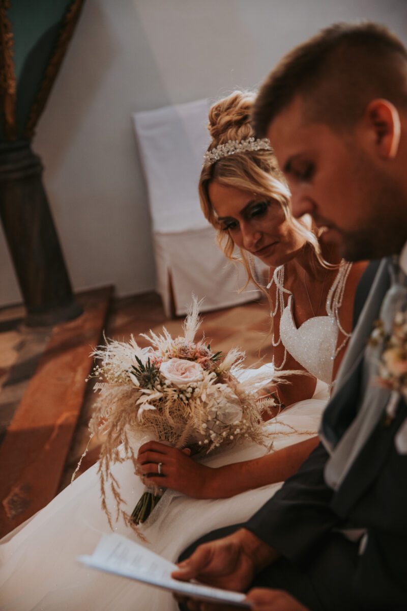 Braut und Bräutigam sitzen eng beieinander und betrachten ein Blatt Papier. Die Braut, die einen Strauß getrockneter Blumen in der Hand hält und ein weißes Kleid mit aufwendigen Spitzendetails und einer Tiara trägt, lächelt herzlich. Der Bräutigam in seinem dunklen Anzug schaut liebevoll zu. Im Hintergrund sind ein Holzboden und eine Wand mit einer dekorativen Säule zu sehen, die ihren intimen Moment perfekt für den Hochzeitsfotografen festhält. - Elis Wedding - Hochzeitsfotograf Frankfurt