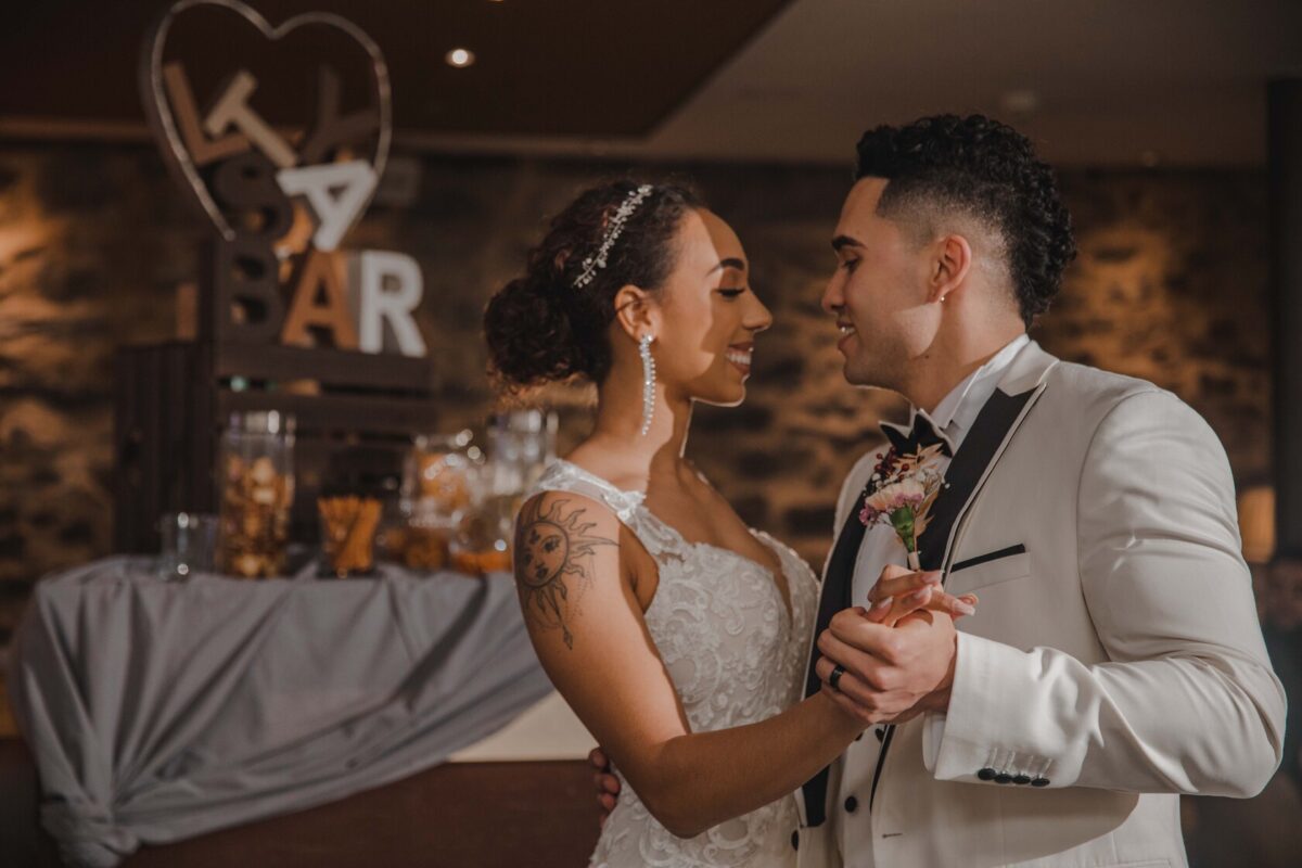 Braut und Bräutigam tanzen bei ihrer Hochzeit. Die Braut, die ein weißes Spitzenkleid und einen Kopfschmuck trägt, lächelt den Bräutigam an, der eine weiße Jacke mit schwarzem Besatz und dazu passende schwarze Hosen trägt. Hinter ihnen steht ein rustikales Holzschild in Herzform mit den Worten „LOVE BAR“ und ein dekorierter Tisch, der den perfekten Moment für ihren Hochzeitsfotografen festhält. - Elis Wedding - Hochzeitsfotograf Frankfurt