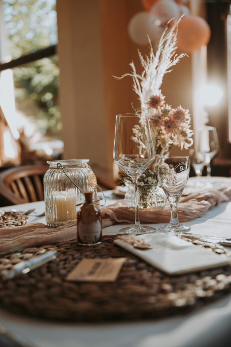 Ein wunderschön gedeckter Tisch für eine Feier, geschmückt mit Weingläsern, einem rustikalen Blumenarrangement mit getrockneten Blumen und Gräsern und einer Kerze im Glas. Das Gedeck umfasst gewebte Tischsets, eine kleine Geschenkflasche und einen zartrosa Tischläufer und fängt die Essenz der Kunst des Hochzeitsfotografen perfekt ein, während Sonnenlicht durch ein nahe gelegenes Fenster fällt. - Elis Wedding - Hochzeitsfotograf Frankfurt