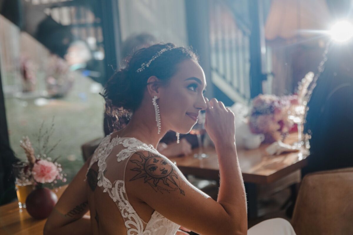 Eine Frau in einem weißen Spitzenkleid mit einem Sonnentattoo auf der Schulter nippt an einem Glas. Sie trägt lange Ohrringe und ein Stirnband. Die Szene ist drinnen mit sanfter Beleuchtung und im Hintergrund sind verschwommene Blumen zu sehen, was deutlich macht, dass sie sich auf einer Veranstaltung wie einer Hochzeitsfeier befinden könnte, perfekt eingefangen vom Hochzeitsfotografen. - Elis Wedding - Hochzeitsfotograf Frankfurt