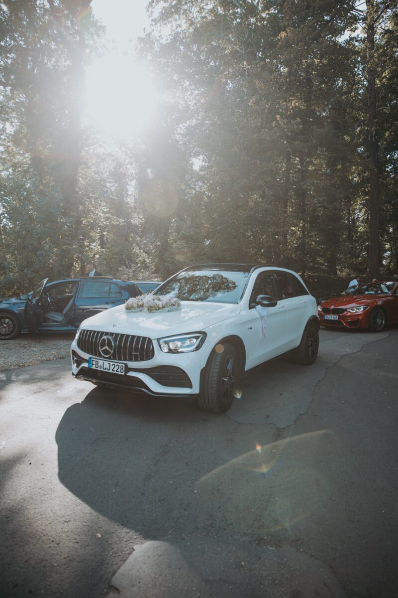 Ein weißer Mercedes-Benz SUV mit Blumendekorationen auf der Motorhaube steht auf einer sonnenbeschienenen Straße, umgeben von Bäumen. Sonnenlicht fällt durch die Äste und erzeugt ein Linsenreflexion, perfekt für ein Fotoshooting. Andere Autos stehen in der Nähe, eine Tür ist offen, was auf eine standesamtliche Trauungsfeier schließen lässt. - Elis Wedding - Hochzeitsfotograf Frankfurt