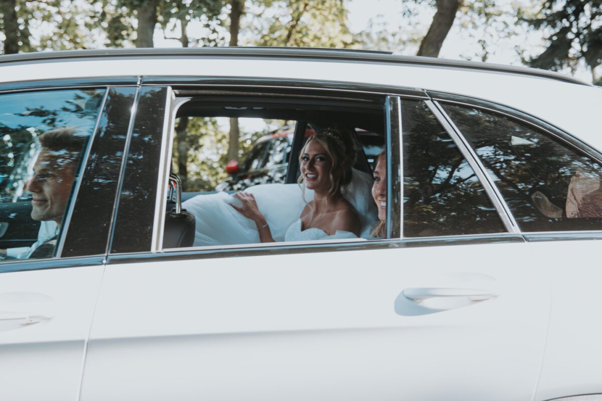Eine Braut in einem weißen Hochzeitskleid sitzt lächelnd in einem weißen Auto. Sie ist durch das offene Fenster zu sehen, in Begleitung einer weiteren Person. Das Foto, das tagsüber mit Bäumen und Sonnenlicht im Hintergrund aufgenommen wurde, fängt einen wunderschönen Moment während ihres standesamtlichen Trauungs-Fotoshootings ein. - Elis Wedding - Hochzeitsfotograf Frankfurt
