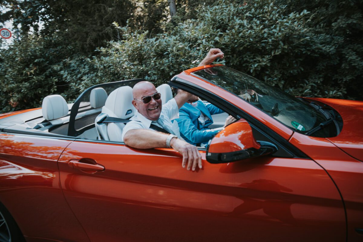 Ein kahlköpfiger Mann mit Sonnenbrille und weißem Hemd fährt in einem roten Cabrio mit offenem Verdeck durch Frankfurt. Er lächelt breit und hat einen Arm auf der Tür ruhend. Auf dem Beifahrersitz sitzt eine weitere Person in blauem Outfit. Saftig grüne Bäume und Blätter bilden eine ideale Kulisse für Fotos. - Elis Wedding - Hochzeitsfotograf Frankfurt