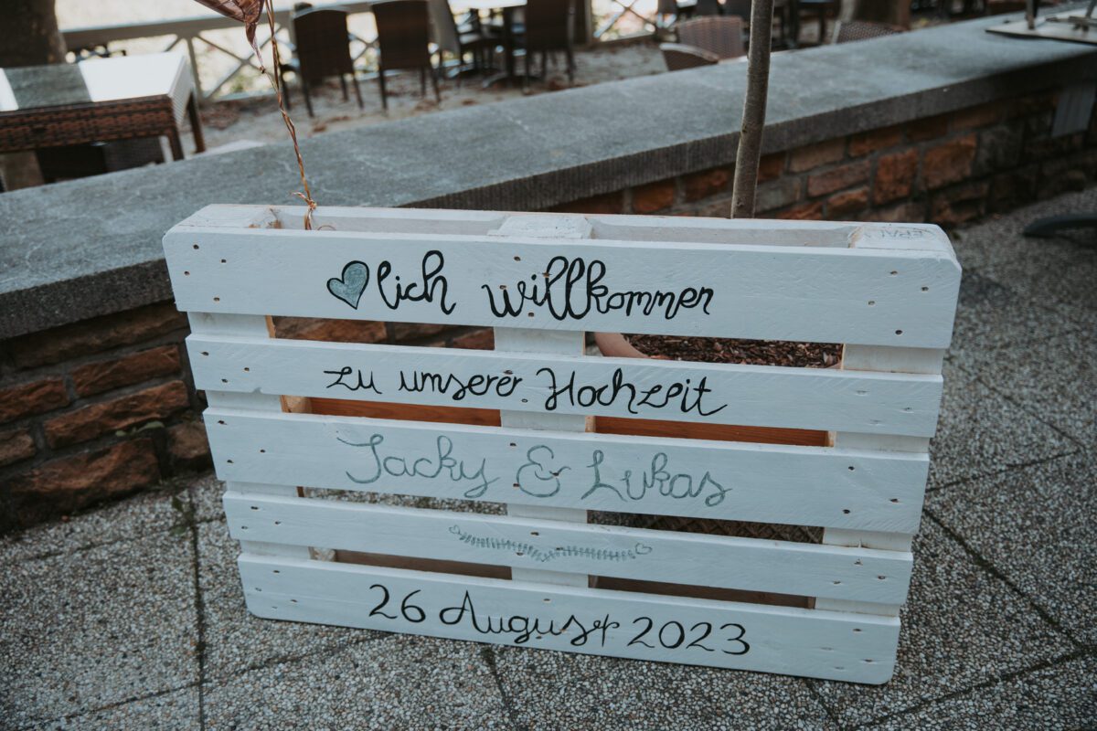 Im Freien steht ein Schild aus einer weißen Holzpalette mit schwarzer und grauer Kalligraphie. Auf dem Schild steht „Herzlich Willkommen zu unserer Hochzeit“, gefolgt von „Jacky & Lukas“ und „26. August 2023“. Das Schild ist perfekt für jeden Hochzeitsfotografen und steht auf einer gepflasterten Fläche in der Nähe eines Holzzauns und eines Sitzbereichs im Freien. - Elis Wedding - Hochzeitsfotograf Frankfurt