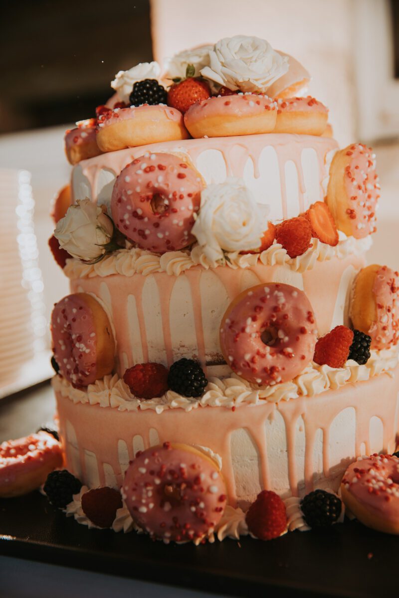 Eine dreistöckige Hochzeitstorte ist mit rosa glasierten Donuts und verschiedenen frischen Beeren, darunter Himbeeren und Brombeeren, geschmückt. Dieses atemberaubende Herzstück, dekoriert mit weißen Rosen und tropfendem weißen Zuckerguss, der jede Etage hinunterfällt, wurde während eines wunderschön beleuchteten Fotoshootings in Frankfurt aufgenommen. - Elis Wedding - Hochzeitsfotograf Frankfurt