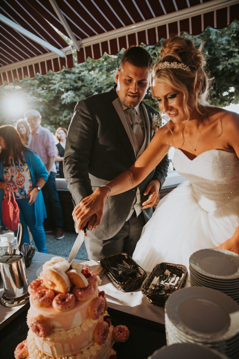 Braut und Bräutigam schneiden während eines fröhlichen Fotoshootings gemeinsam ihre Hochzeitstorte an. Die Braut trägt ein weißes trägerloses Kleid und ihre Haare sind elegant hochgesteckt, während der Bräutigam einen schicken Anzug trägt. Die Torte ist mit rosa Donuts dekoriert und die Gäste sind unter einem Vordach im Freien zu sehen. - Elis Wedding - Hochzeitsfotograf Frankfurt