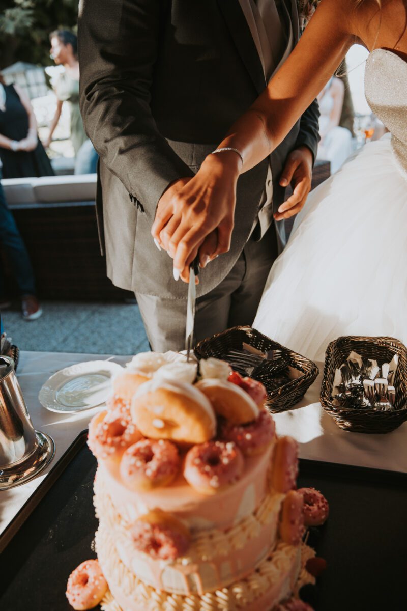 Braut und Bräutigam in Hochzeitskleidung schneiden eine mehrstöckige Torte an, die mit rosa glasierten Donuts geschmückt ist. Die Braut trägt ein weißes schulterfreies Kleid und der Bräutigam einen grauen Anzug. Ihre Hände liegen zusammen auf dem Messer, im Hintergrund sind Gäste und Geschirr bei ihrer Standesamtlichen Trauung in Frankfurt zu sehen. - Elis Wedding - Hochzeitsfotograf Frankfurt