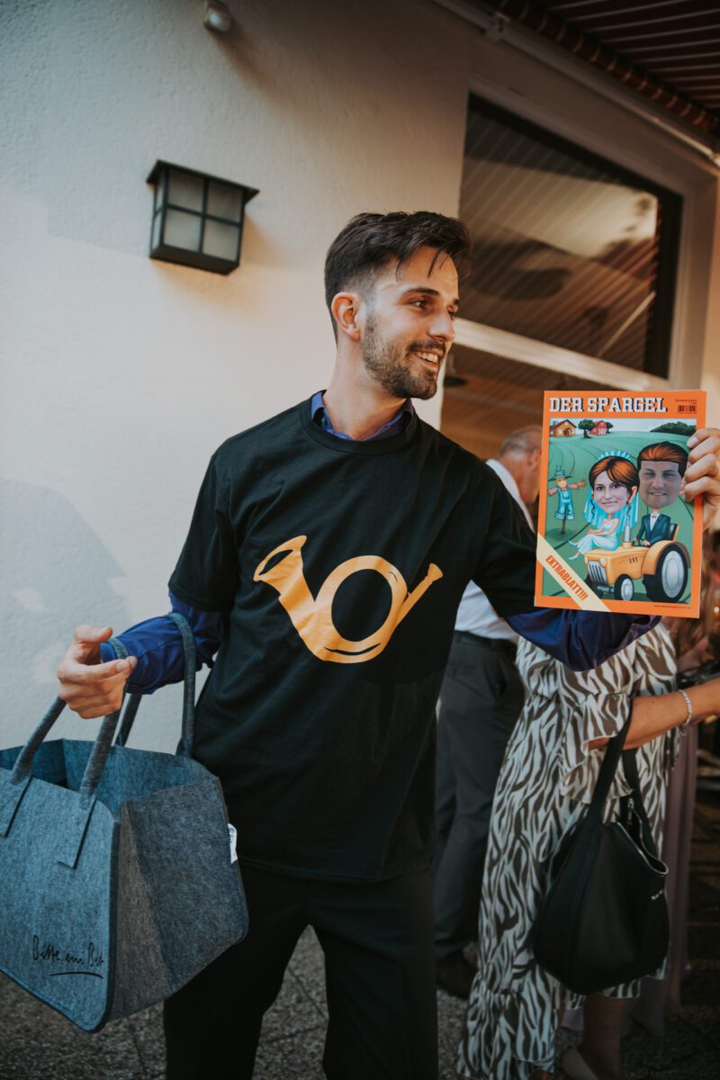 Ein Mann in einem schwarzen T-Shirt mit gelbem Horn-Motiv hält in der einen Hand eine Einkaufstüte und in der anderen eine Zeitschrift mit dem Titel „Der Spiegel“. Auf dem Cover der Zeitschrift sind animierte Figuren zu sehen. Im Hintergrund sind eine weitere Person und ein Gebäudeteil mit einer an der Wand hängenden Laterne zu sehen, was eine ideale Kulisse für Fotografien darstellt. - Elis Wedding - Hochzeitsfotograf Frankfurt