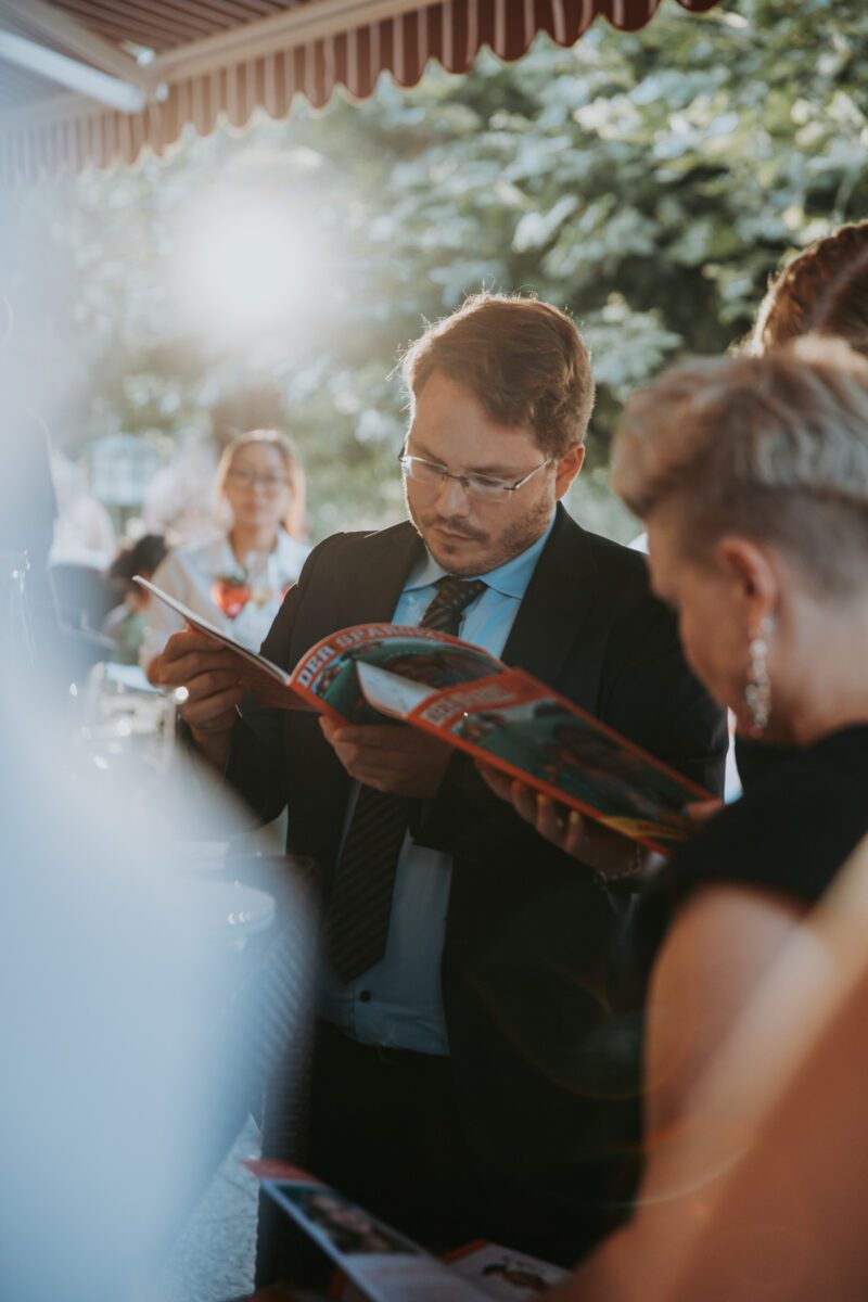 Ein Mann in Anzug und Krawatte liest eine Zeitschrift, während er draußen unter einem Vordach steht und sich möglicherweise einen Moment Zeit für eine standesamtliche Trauung nimmt. Er trägt eine Brille und hat einen konzentrierten Gesichtsausdruck. Im Hintergrund scheint Sonnenlicht, um ihn herum sind teilweise Grünflächen und andere Menschen zu sehen. Eine Frau mit kurzen Haaren und Ohrringen steht in der Nähe und blickt abgewandt. - Elis Wedding - Hochzeitsfotograf Frankfurt
