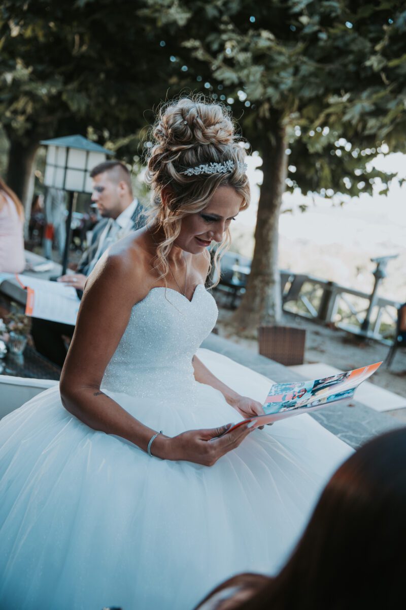 Eine Braut mit einer aufwendigen Hochsteckfrisur und einer funkelnden Tiara sitzt im Freien und lächelt, während sie eine bunte Zeitschrift oder ein Fotobuch betrachtet. Sie trägt ein weißes, trägerloses Brautkleid mit einem taillierten Mieder und einem voluminösen Rock. Im Hintergrund sind Bäume und weiches, natürliches Licht zu sehen, perfekt für Hochzeitsfotografen. - Elis Wedding - Hochzeitsfotograf Frankfurt