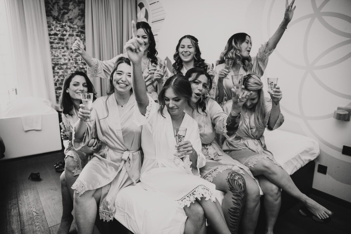 Eine Gruppe von Frauen in Bademänteln sitzt und steht um ein Bett, stößt mit den Gläsern an und lächelt. Das Bild fängt einen feierlichen Moment ein, möglicherweise eine Hochzeitsgesellschaft vor der Standesamtlichen Trauung in Frankfurt. Der Hintergrund zeigt eine gemusterte Wand und ein Fenster mit Vorhängen, wodurch eine lebhafte Szene entsteht, an der alle freudig teilnehmen. - Elis Wedding - Hochzeitsfotograf Frankfurt