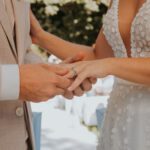 Eine Person im beigen Jackett steckt einer anderen Person einen Ring an den Finger. Sie trägt ein weißes Kleid mit tiefem Ausschnitt und Blumenmuster. Ihre Umgebung ist sanft verschwommen, mit Andeutungen von Bäumen und einer Außenkulisse, die an ein Fotoshooting für eine Hochzeitszeremonie in Frankfurt denken lassen. - Elis Wedding - Hochzeitsfotograf Frankfurt