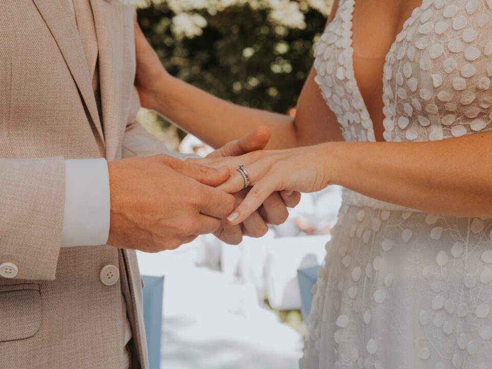 Eine Person im beigen Jackett steckt einer anderen Person einen Ring an den Finger. Sie trägt ein weißes Kleid mit tiefem Ausschnitt und Blumenmuster. Ihre Umgebung ist sanft verschwommen, mit Andeutungen von Bäumen und einer Außenkulisse, die an ein Fotoshooting für eine Hochzeitszeremonie in Frankfurt denken lassen. - Elis Wedding - Hochzeitsfotograf Frankfurt