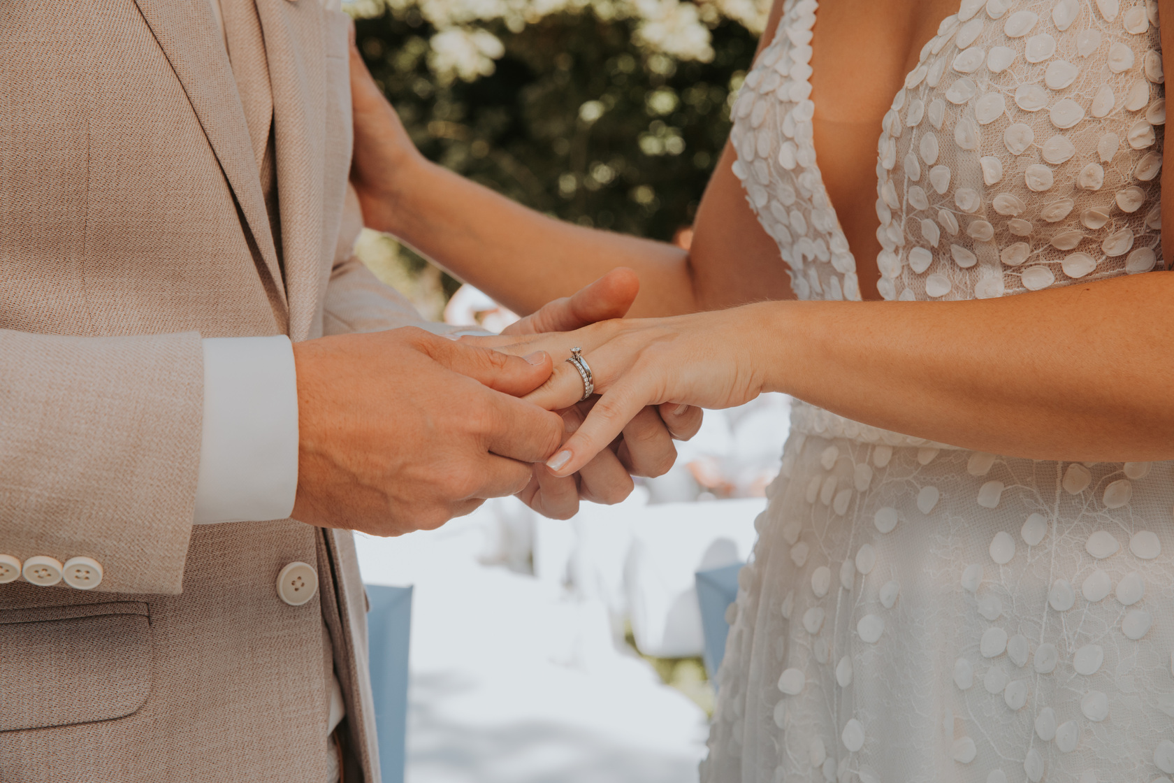 Eine Person im beigen Jackett steckt einer anderen Person einen Ring an den Finger. Sie trägt ein weißes Kleid mit tiefem Ausschnitt und Blumenmuster. Ihre Umgebung ist sanft verschwommen, mit Andeutungen von Bäumen und einer Außenkulisse, die an ein Fotoshooting für eine Hochzeitszeremonie in Frankfurt denken lassen. - Elis Wedding - Hochzeitsfotograf Frankfurt