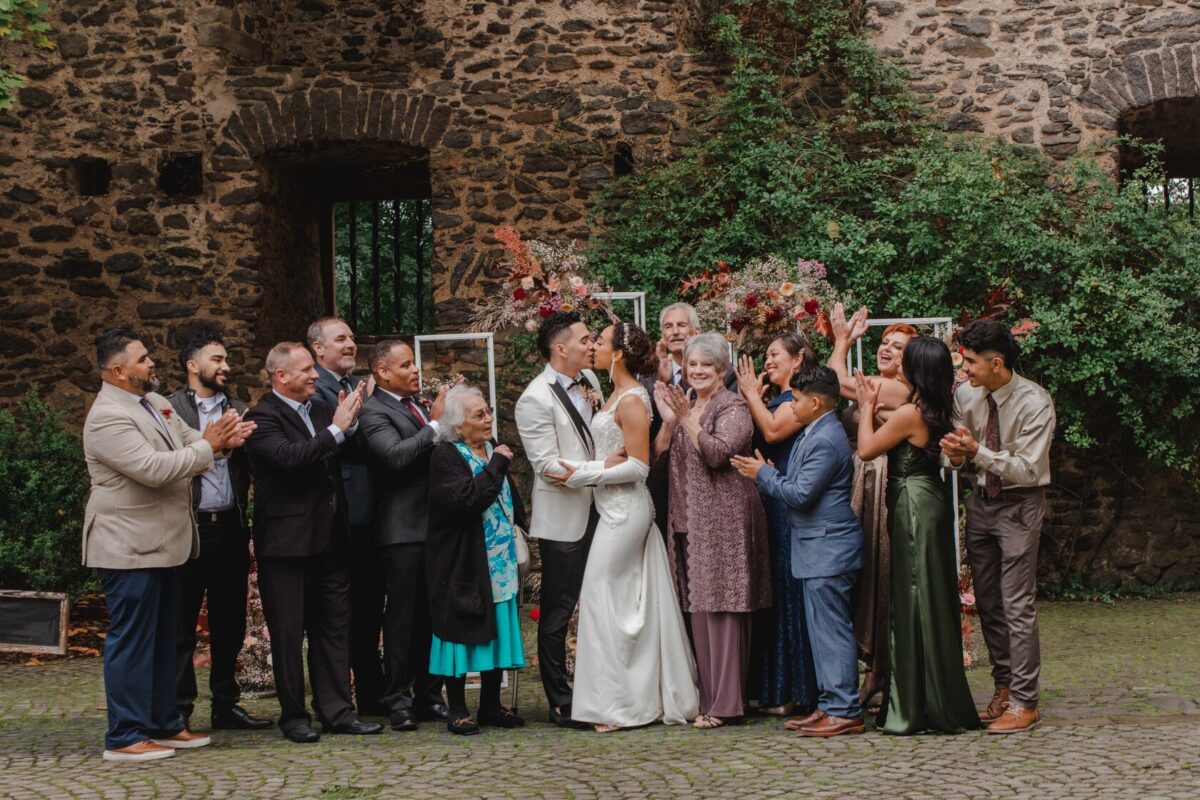 Eine fröhliche Hochzeitsszene zeigt Braut und Bräutigam, die sich während ihrer standesamtlichen Trauung in der Mitte küssen. Sie sind von einer applaudierenden Gruppe von Familie und Freunden umgeben. Der Hintergrund ist eine rustikale Steinmauer, die mit üppigem Grün und Blumenarrangements geschmückt ist und den perfekten Moment für die Fotografie einfängt. - Elis Wedding - Hochzeitsfotograf Frankfurt