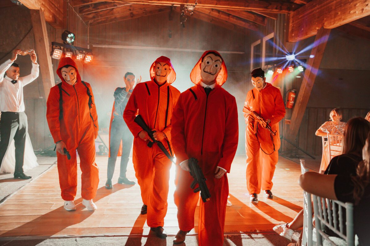 Fünf Personen in orangefarbenen Overalls und Dali-Masken stehen in einer Holzhalle mit dramatischer Beleuchtung und bereiten sich wahrscheinlich auf ein exzentrisches Fotoshooting vor. Zwei von ihnen halten Requisitenwaffen, während ein anderer in weißem Hemd und Fliege im Hintergrund tanzt. Rauch und bunte Lichter verstärken die festliche, theatralische Atmosphäre, während sitzende Gäste zuschauen. - Elis Wedding - Hochzeitsfotograf Frankfurt