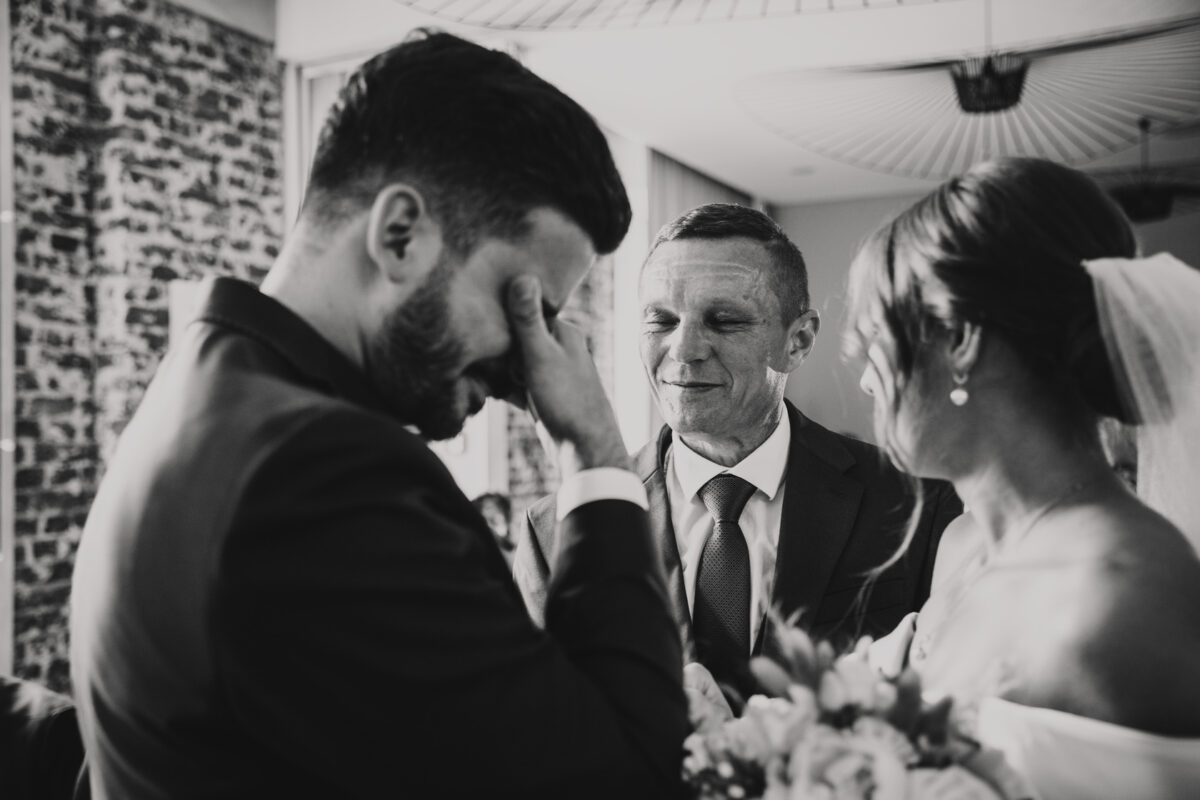 Eine Schwarz-Weiß-Fotografie fängt einen emotionalen Hochzeitsmoment ein. Der Bräutigam ist von seinen Gefühlen überwältigt und bedeckt sein Gesicht mit einer Hand. Neben ihm steht ein älterer Mann, der warmherzig lächelt. Die Braut, mit einem Schleier geschmückt und einem Blumenstrauß in der Hand, steht ihnen gegenüber und fängt den intimen Austausch durch die Linse eines erfahrenen Hochzeitsfotografen ein. - Elis Wedding - Hochzeitsfotograf Frankfurt