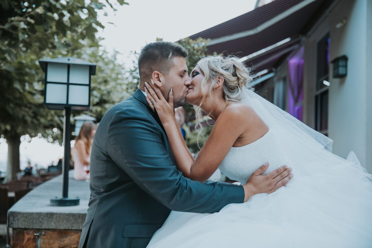 Ein frisch vermähltes Paar küsst sich draußen zärtlich. Der Hochzeitsfotograf fotografiert den Bräutigam in einem dunklen Anzug, wie er zärtlich das Gesicht der Braut hält. In einem weißen trägerlosen Kleid und Schleier umarmt sie ihn neben einem Steinvorsprung mit einem Laternenpfahl vor einem Gebäude mit schrägem Dach. - Elis Wedding - Hochzeitsfotograf Frankfurt