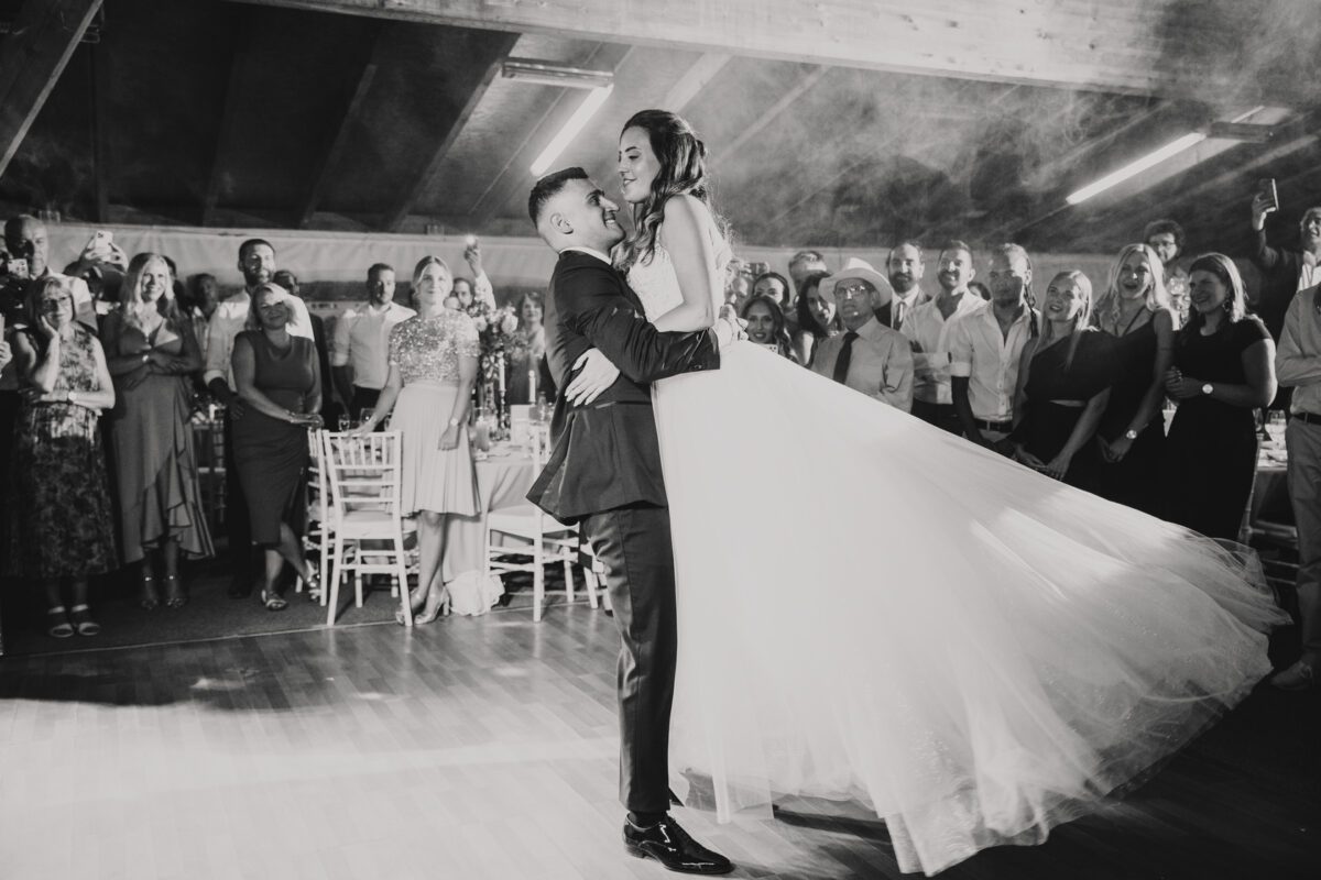 Ein Hochzeitsfotograf fängt einen magischen Moment ein, als der Bräutigam die Braut hochhebt und auf der Tanzfläche in einem geräumigen, schwach beleuchteten Veranstaltungsort in Frankfurt wirbelt. Die Braut in einem fließenden weißen Kleid lächelt freudig. Elegante Gäste beobachten den Tanz sichtlich bewegt. Sanftes Licht und Holzboden tragen zur warmen Atmosphäre der Feier bei. - Elis Wedding - Hochzeitsfotograf Frankfurt