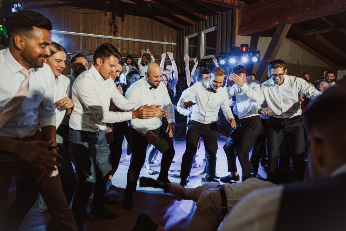 Eine lebhafte Gruppe von Männern, alle in weißen Hemden und dunklen Hosen, tanzt begeistert bei einer Indoor-Veranstaltung. Einige haben ihre Hände in die Luft gestreckt. Alle scheinen Spaß zu haben, und in der Mitte tanzt ein Mann energisch auf dem Boden. Die Kulisse besteht aus Holzwänden und -balken – eine perfekte Szene für einen aufstrebenden Hochzeitsfotografen. - Elis Wedding - Hochzeitsfotograf Frankfurt