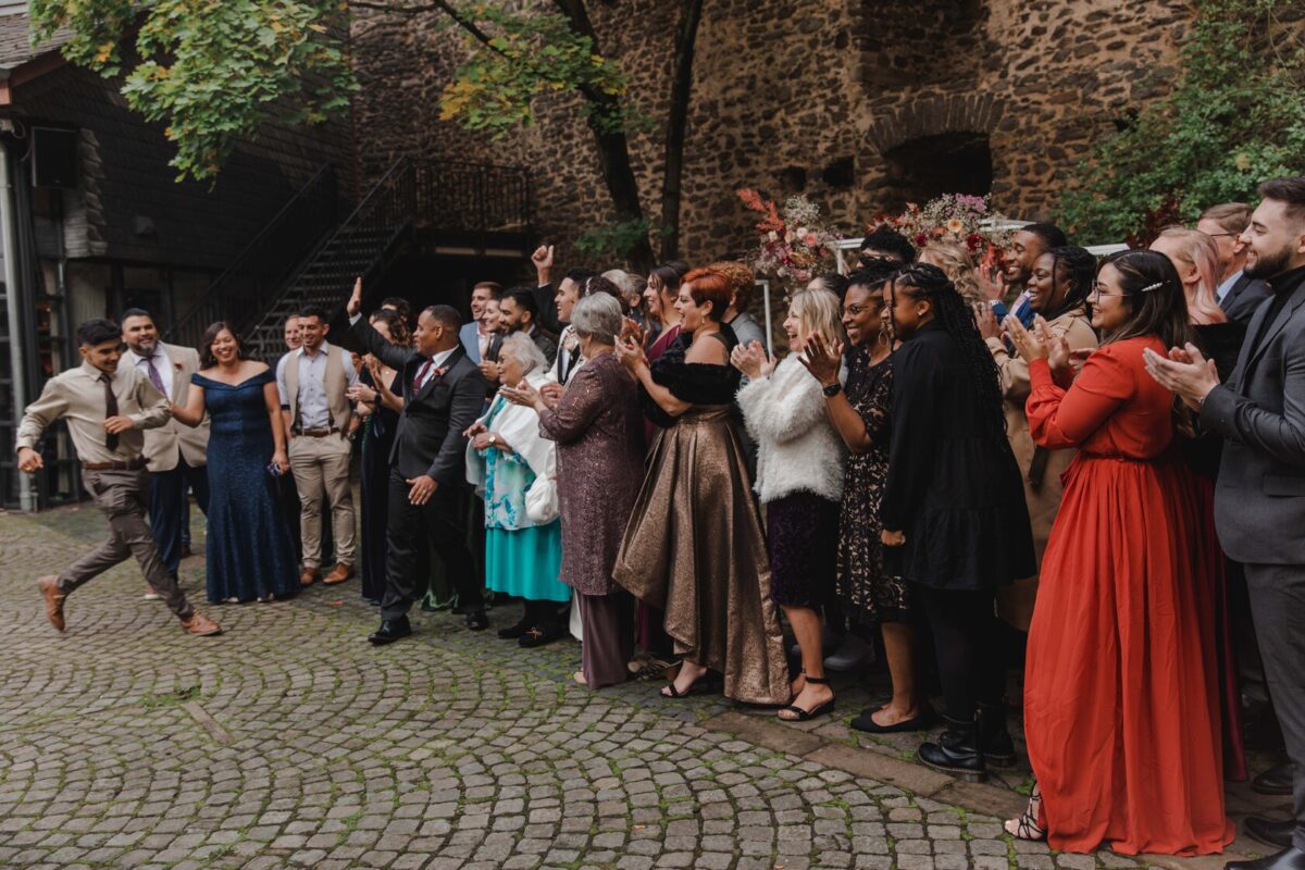 Eine fröhliche Gruppe von Hochzeitsgästen applaudiert einem Mann im beigen Anzug, der tanzt und Leuten High Fives gibt. Die Gäste, in formeller Kleidung gekleidet, stehen auf einem Kopfsteinpflasterhof vor einer Kulisse aus rustikalen Steinhäusern und Bäumen in Frankfurt und schaffen eine festliche und feierliche Atmosphäre. - Elis Wedding - Hochzeitsfotograf Frankfurt