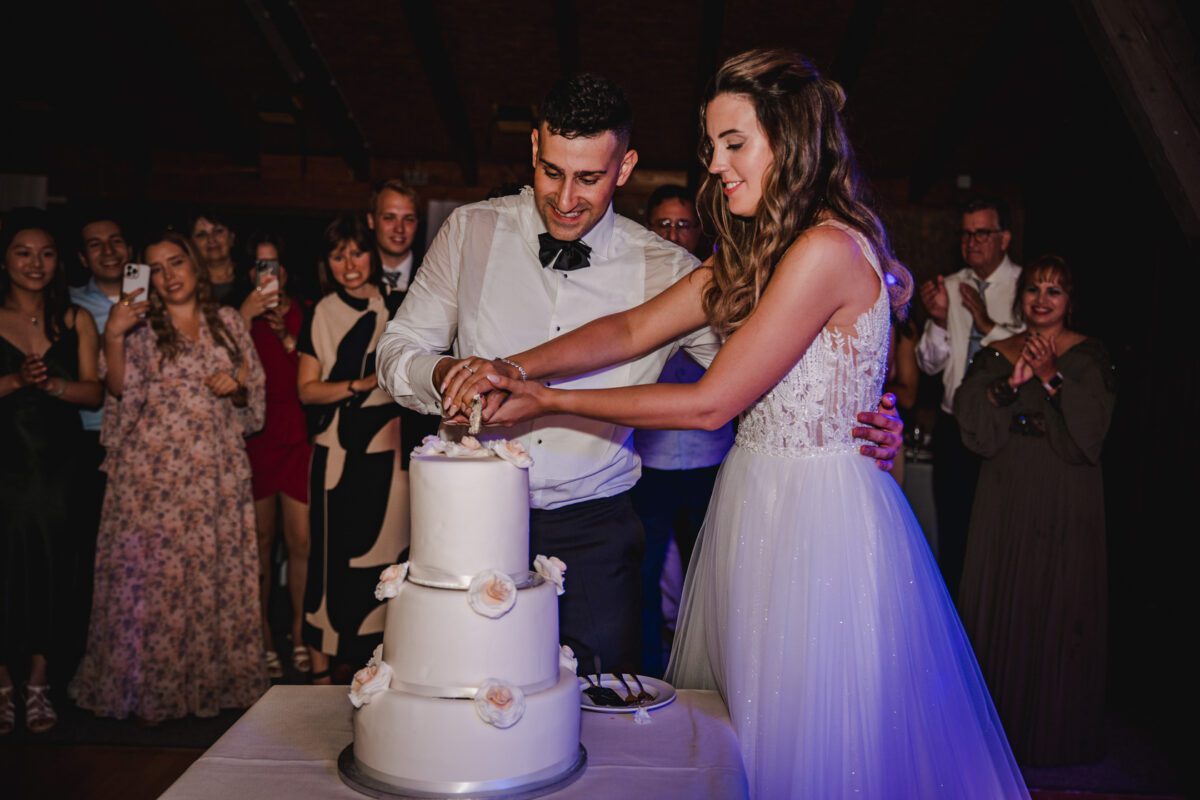 Braut und Bräutigam schneiden gemeinsam eine dreistöckige weiße Hochzeitstorte an. Die Braut trägt ein weißes Kleid mit strukturiertem Mieder und der Bräutigam ein weißes Hemd mit schwarzer Fliege. Gäste in Abendgarderobe umringen sie, lächeln und machen Fotos, während der Hochzeitsfotograf diese kostbaren Momente im Haus festhält. - Elis Wedding - Hochzeitsfotograf Frankfurt