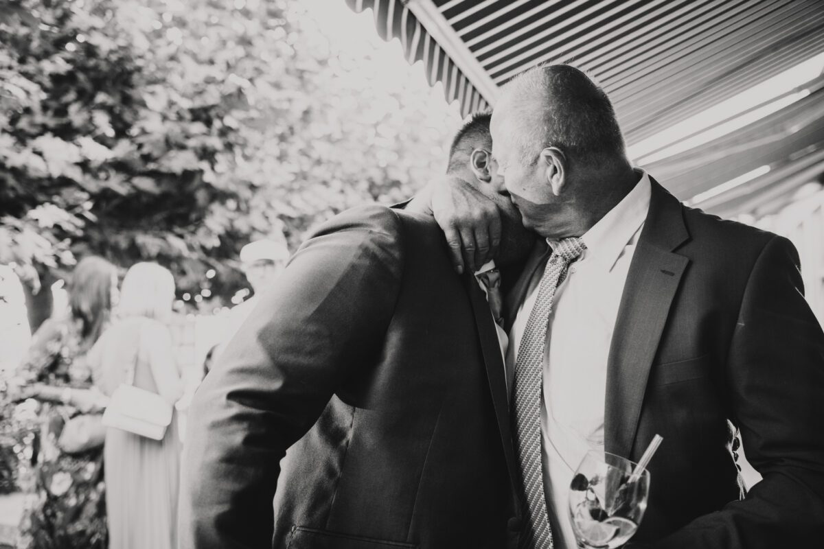 Schwarz-Weiß-Fotografie von zwei Männern in Anzügen, die sich herzlich umarmen. Ein Mann hat seinen Arm um die Schulter des anderen gelegt, ihre Gesichter liegen nah beieinander, was eine tiefe emotionale Bindung suggeriert. Im Hintergrund sind mehrere Menschen versammelt, wodurch eine leicht verschwommene, belebte Außenkulisse entsteht, die an Frankfurt erinnert. - Elis Wedding - Hochzeitsfotograf Frankfurt