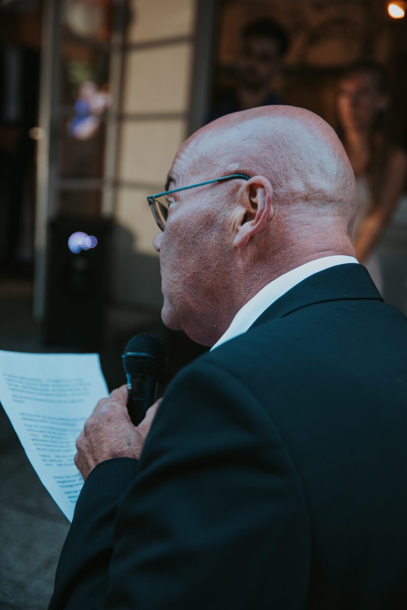 Ein kahlköpfiger Mann mit Brille und schwarzem Anzug spricht in ein Mikrofon und hält dabei ein Stück Papier mit aufgedrucktem Text in der Hand. Er scheint eine Ansprache zu halten, möglicherweise bei einer standesamtlichen Trauung. Durch den leicht verschwommenen Hintergrund in dem schwach beleuchteten Bereich sind Menschen zu sehen. - Elis Wedding - Hochzeitsfotograf Frankfurt