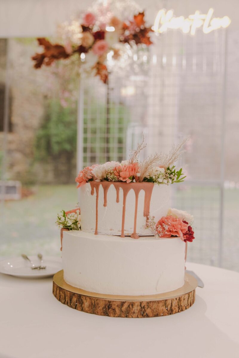 Eine zweistöckige Torte, verziert mit weißem Zuckerguss und einer hellrosa Tropfglasur, liegt auf einer Holzplatte. Die oberste Etage ist mit Blumen in Pfirsich-, Rosa- und Weißtönen dekoriert. Im unscharfen Hintergrund sind herbstliche Dekorationen und ein neonfarbenes „Love“-Schild zu sehen, perfekt für ein unvergessliches Fotoshooting während einer standesamtlichen Trauung.
 - Elis Wedding - Hochzeitsfotograf Frankfurt