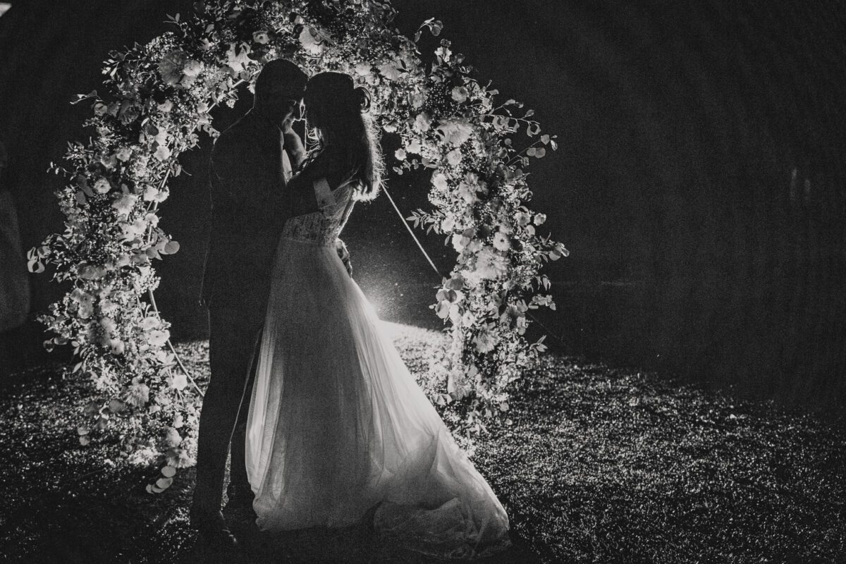 Ein Schwarzweißfoto von Braut und Bräutigam in Frankfurt, die eng beieinander unter einem runden Blumenbogen stehen. Das Paar ist von hinten beleuchtet, wodurch ein romantischer Silhouetteneffekt entsteht. Die Braut trägt ein langes, fließendes Kleid und der Bräutigam einen Anzug. Der Hintergrund ist dunkel und betont den intimen Moment ihrer standesamtlichen Trauung. - Elis Wedding - Hochzeitsfotograf Frankfurt
