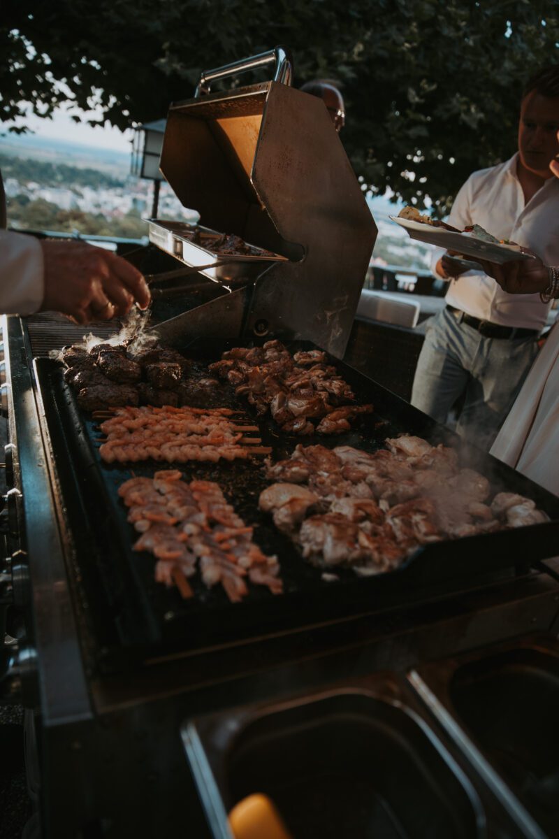 Ein Grill voller verschiedener Fleischsorten, darunter Spieße und Steaks, wird im Freien gegrillt. Eine Person in einem weißen Hemd ist zu sehen, wie sie das Fleisch mit einer Zange wendet, während andere herumstehen und einer einen Teller hält. Die Szene fängt ein informelles Wiedersehen mit einem belaubten Baum im Hintergrund und einer bewölkten Stadtansicht ein, perfekt für ein spontanes Fotoshooting. - Elis Wedding - Hochzeitsfotograf Frankfurt