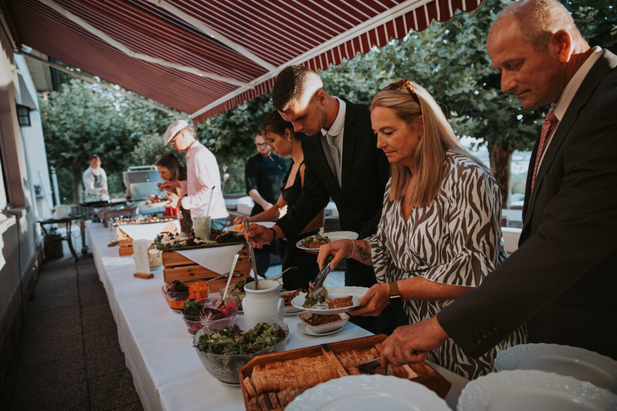 Eine Gruppe formell gekleideter Menschen bedient sich an einem Buffettisch im Freien bei einer Standesamtlichen Trauung in Frankfurt. Der Tisch ist mit einem weißen Tuch bedeckt und bietet eine Auswahl an Gerichten, darunter Salate, Brot und andere Vorspeisen. Im Hintergrund sind rot-weiß gestreifte Markisen und Bäume zu sehen. - Elis Wedding - Hochzeitsfotograf Frankfurt