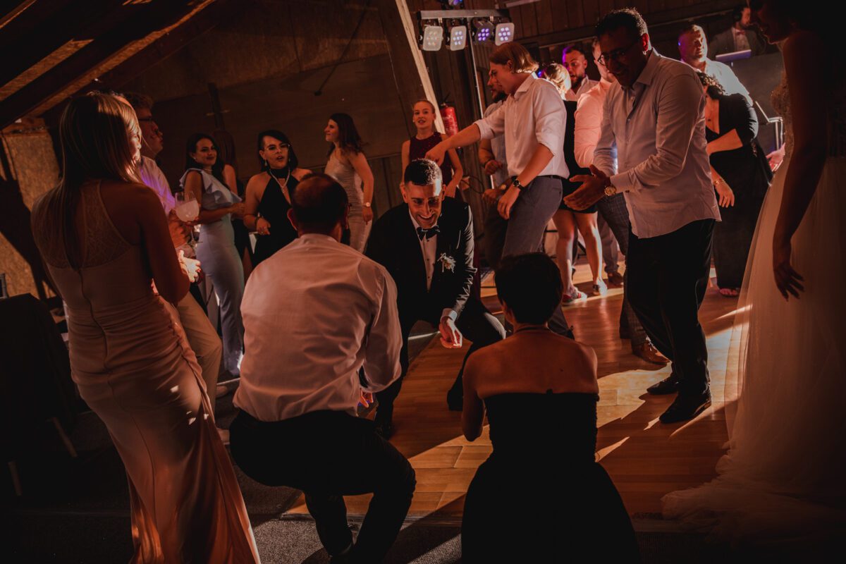 Eine Gruppe formell gekleideter Menschen tanzt energisch auf einer Indoor-Party mit gedämpftem, warmem Licht. Der Mann in der Mitte, in dunklem Anzug und Fliege, hockt tief. Um ihn herum sind Männer und Frauen in Kleidern und Anzügen, alle in den lebhaften Tanz vertieft – vielleicht feiern sie nach einer Standesamtlichen Trauung in Frankfurt. - Elis Wedding - Hochzeitsfotograf Frankfurt