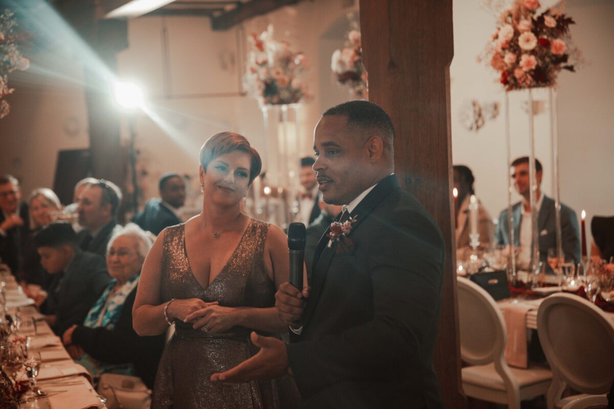 Bei einer formellen Veranstaltung hält ein Mann im Anzug mit einer Boutonniere ein Mikrofon und scheint eine Rede zu halten. Eine Frau in einem funkelnden Kleid steht lächelnd neben ihm. Sie sind von Gästen umgeben, die an elegant dekorierten Tischen mit Blumenschmuck und Kerzen sitzen, was eine festliche oder feierliche Atmosphäre suggeriert, eingefangen von einem Hochzeitsfotografen in Frankfurt. - Elis Wedding - Hochzeitsfotograf Frankfurt