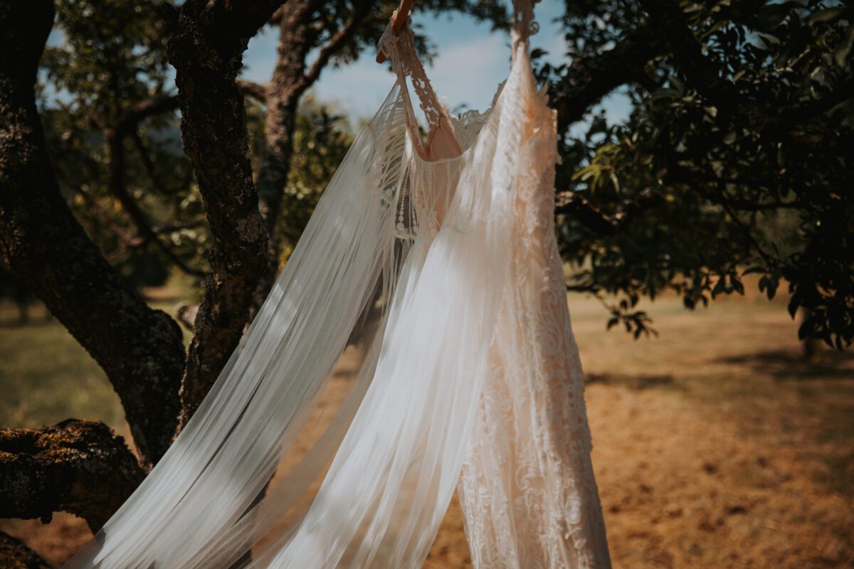 Ein weißes, aufwendig gestaltetes Hochzeitskleid hängt an einem Ast. Das Kleid ist mit Spitzendetails und einer fließenden Schleppe aus Tüll versehen. Sonnenlicht fällt durch die Blätter und wirft gesprenkeltes Licht auf den Stoff. Der von einem Hochzeitsfotografen aufgenommene Hintergrund zeigt eine Rasenfläche mit Bäumen, was eine Szenerie im Freien suggeriert. - Elis Wedding - Hochzeitsfotograf Frankfurt