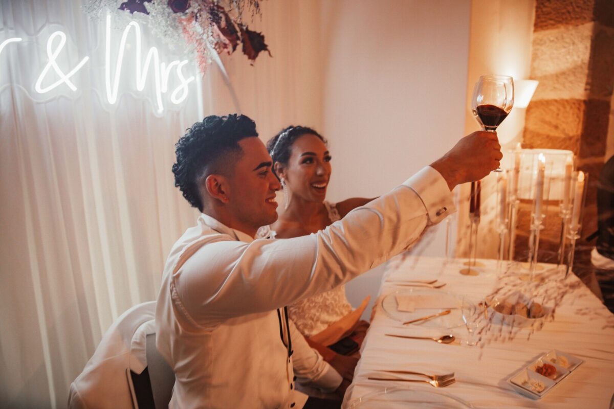 Ein Paar in Hochzeitskleidung sitzt an einem Banketttisch. Der Bräutigam, in einem weißen Hemd, erhebt ein Glas Rotwein zum Anstoßen, während die Braut in ihrem weißen Spitzenkleid neben ihm lächelt. Aufgenommen von ihrem Hochzeitsfotografen aus Frankfurt feiern sie ihre standesamtliche Trauung unter einem Neonschild mit der Aufschrift „Mr & Mrs“. Der Tisch ist elegant gedeckt - Elis Wedding - Hochzeitsfotograf Frankfurt