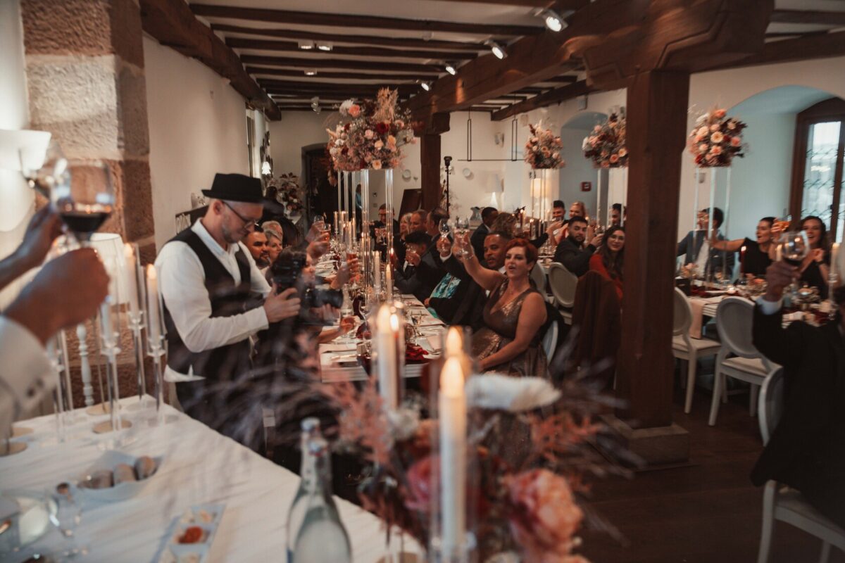 Ein lebhafter Bankettsaal voller Gäste, die an langen Tischen sitzen, die mit Blumenschmuck und Kerzen geschmückt sind. Die Leute erheben ihre Gläser zum Toast. Ein Hochzeitsfotograf in schwarzem Hut und Weste macht auf der linken Seite ein Foto, während andere lächeln und sich unterhalten. Die Atmosphäre ist festlich und warm. - Elis Wedding - Hochzeitsfotograf Frankfurt
