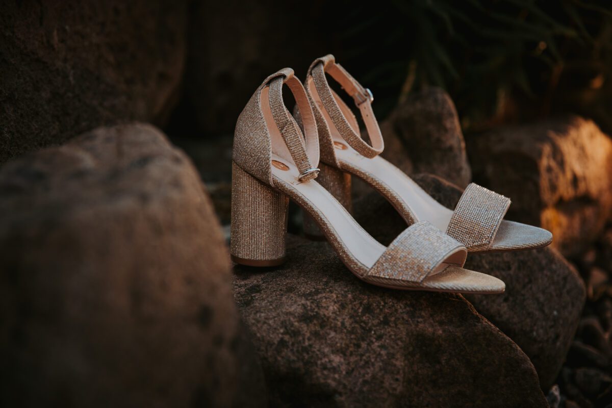 Ein Paar eleganter, offener High-Heel-Sandalen mit Knöchelriemen steht auf rauen, strukturierten Steinen. Die Schuhe haben eine schimmernde, metallisch beige Farbe, reflektieren das Licht subtil, haben klobige Absätze und ein anspruchsvolles, minimalistisches Design. Weiches, natürliches Licht wertet die Szene auf – perfekt für einen Hochzeitsfotografen in Frankfurt. - Elis Wedding - Hochzeitsfotograf Frankfurt