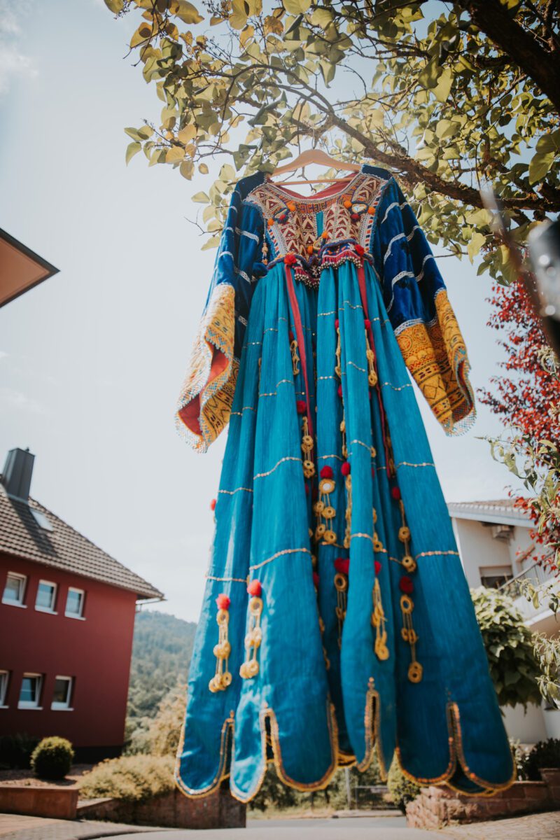 Ein farbenfrohes traditionelles Kleid hängt sanft wiegend an einem Ast. Das Kleidungsstück, perfekt für ein Fotoshooting, ist in leuchtendem Türkis gehalten und mit aufwendigen goldenen, roten und gelben Details wie Mustern und Pompons verziert. Der Hintergrund besteht aus einem klaren blauen Himmel, Bäumen und Wohnhäusern – eines in Rot und eines in Weiß. - Elis Wedding - Hochzeitsfotograf Frankfurt