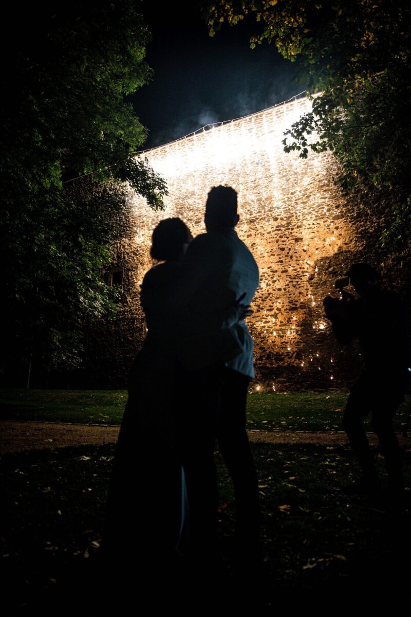 Im Vordergrund steht ein Paar vor einer Steinmauer, die mit herabhängenden Lichterketten geschmückt ist, die sich gegen den Nachthimmel abheben. Die Silhouetten der Bäume rahmen die Szene ein. Rechts hält ein Fotograf den Moment dieses ruhigen und romantischen Fotoshootings in Frankfurt fest. - Elis Wedding - Hochzeitsfotograf Frankfurt