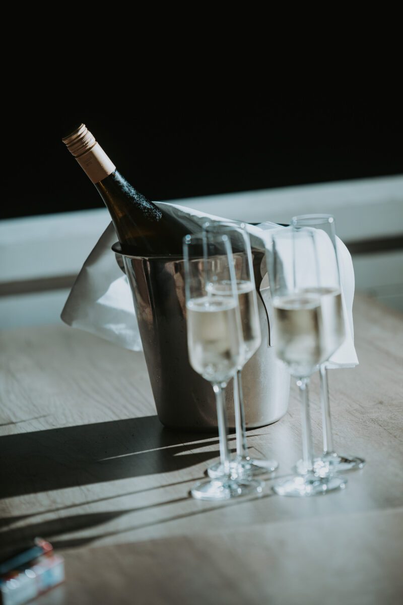 Zwei mit Sekt gefüllte Champagnergläser stehen auf einem Holztisch. Dahinter steht eine Flasche gekühlten Weins in einem silbernen Eiskübel, teilweise bedeckt von einer weißen Stoffserviette. Das Licht wirft Schatten auf den Tisch und schafft eine mondäne und festliche Atmosphäre – perfekt für Ihren Hochzeitsfotografen. - Elis Wedding - Hochzeitsfotograf Frankfurt