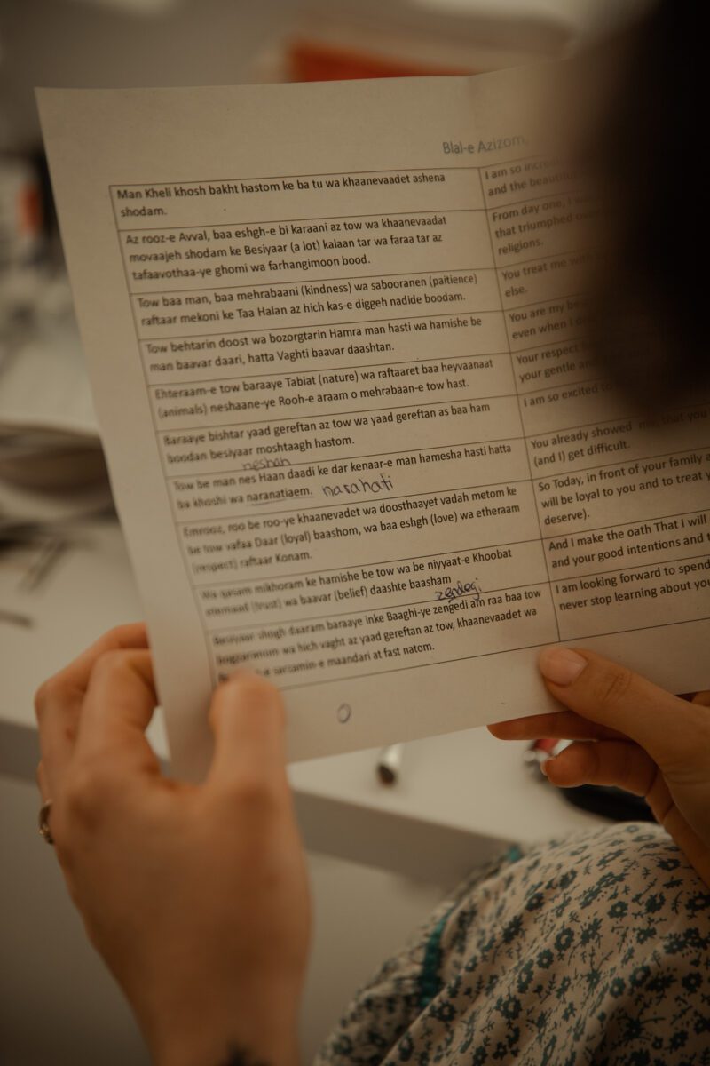 Eine Person hält ein Blatt Papier mit mehreren Textabsätzen in einer nicht-lateinischen Schrift. Ihre mit Nagellack verzierten Hände sind sichtbar. Oben auf dem Papier steht „Blade Azizam“ und der Text ist in einem Tabellenformat angeordnet. Im unscharfen Hintergrund, der an ein Fotoshooting in Frankfurt erinnert, steht ein Schreibtisch mit verschiedenen Objekten. - Elis Wedding - Hochzeitsfotograf Frankfurt